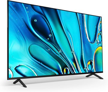 Černý televizor s plochou obrazovkou s tenkým kovovým rámem, lesklý panel pokrývající téměř celý přední povrch, dva černé nohy ve tvaru V na každém konci, pohled z levého předního tříčtvrtečního úhlu