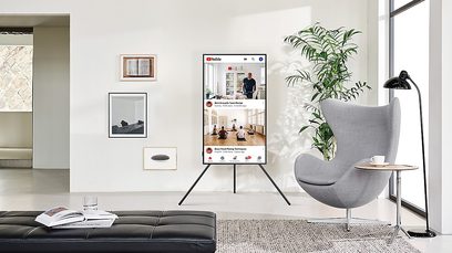 Vægmonteret Samsung TV VG-ARAB22STDXC i en moderne stue med gråt sofa, lænestol, grønne planter og stort vindue