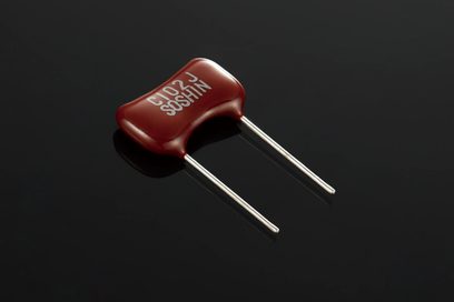 Condensador rojo de cerámica con la inscripción "F 027 SOSHIN" y dos patillas metálicas, vista cercana sobre fondo negro