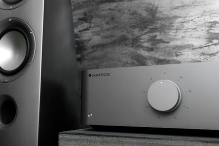 Amplificator audio gri cu buton rotativ, pe o suprafață gri, lângă un difuzor negru, textură lemn pe fundal