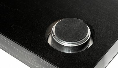 Cirkulær metalvibrationsabsorbator med børstet overflade på mørk træbase, set oppefra i nærbillede