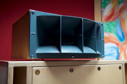 Søjlehøjttaler i mørkt træ og metal, 3 sorte højttalere synlige bag et metalgitter, placeret på et møbel i lyst træ