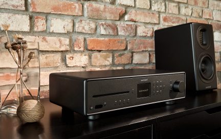 Černý hi-fi systém položený na dřevěném nábytku před červenou cihlovou zdí. Váza s uschlými větvemi vlevo.