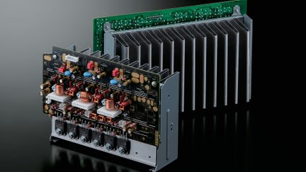 A complex printed circuit board with electronic components, copper coils, a metal heat sink, and a green board in the background