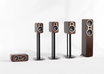 Set of five hi-fi speakers with brown wood finish and black cones, front view, including a horizontal center speaker with three drivers, two identical compact speakers mounted on black metal stands, one compact speaker placed on the floor, and a taller floorstanding speaker equipped with two woofers and a central tweeter