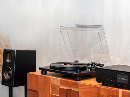 Altavoz negro rectangular, tocadiscos negro con tapa transparente abierta, reproductor negro, mueble de madera, pared clara