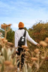 The photo shows a person from behind walking in nature, wearing a backpack and an orange beanie, on a beautiful sunny day