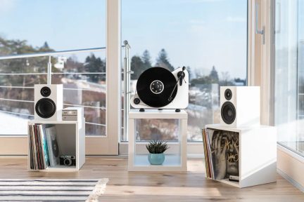 Balta vinilinė plokštelė su juodu disku, 2 baltos kolonėlės, 2 balti baldai su vinilinėmis plokštelėmis, fotoaparatas, mėlynas vazonas, medinės grindys.