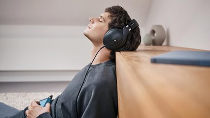 Persona seduta di profilo, appoggiata a una superficie di legno, indossa cuffie audio nere e tiene uno smartphone in mano