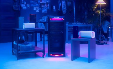Vista frontale di una torre audio nera in plastica con griglia metallica e illuminazione LED rosa-blu, circondata da cinque diffusori portatili rettangolari in plastica nera, bianca e blu dotati di aloni luminosi, disposti su piccoli mobili in una stanza con illuminazione blu.
