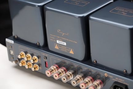 Nærbillede af bagsiden i skrå vinkel af en integreret rørforstærker i blågrå metal, rektangulære transformatorhætter, guldbelagte RCA-stik og gennemsigtige højttalerterminaler