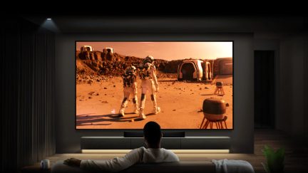 Pantalla rectangular que muestra una escena marciana con 2 astronautas, módulos, rocas, mobiliario, vista desde un sofá.