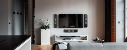 Moderne stue set forfra med sort fladskærms-tv monteret på væggen mellem to sorte matte højttalere med sølvfarvede metalringe, tilhørende centerhøjttaler nedenunder, hvid lakkeret væghylde med apparater og bøger, mat hvid blomsterpotte med orkidé til venstre og grå stofsofa i forgrunden