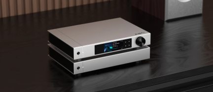 Rechthoekig hi-fi audioapparaat van zilverkleurig geborsteld aluminium, driekwart vooraanzicht van rechts, voorzijde met kleurenscherm, vijf zwarte knoppen, grote zwarte draaiknop rechts, twee hoofdtelefoonaansluitingen links, zijkanten voorzien van zwarte koelribben