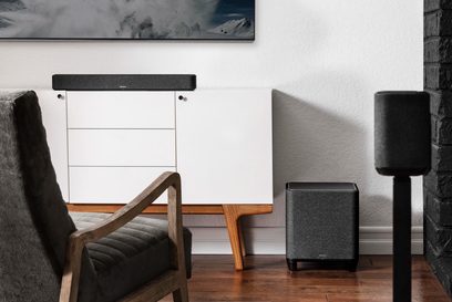 Denon Home Subwoofer svart basförstärkare, svart kubisk högtalare, grå fåtölj, parkettgolv, vit vägg, tavla med snöklädda berg