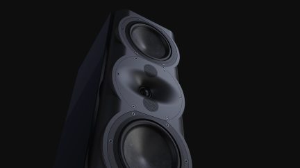 Enceinte noire vue en angle, 3 haut-parleurs circulaires visibles, 2 grilles métalliques centrales, surface lisse, fond noir