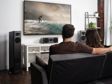 Due persone sedute su un divano grigio, schermo che mostra un surfista, altoparlanti neri, mobile bianco con oggetti decorativi.