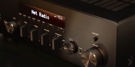 Črni audio ojačevalnik iz četkanega kovine, zaslon 'Net Radio', velik krmilnik glasnosti in izbirniki, pogled od spred tri četrtine