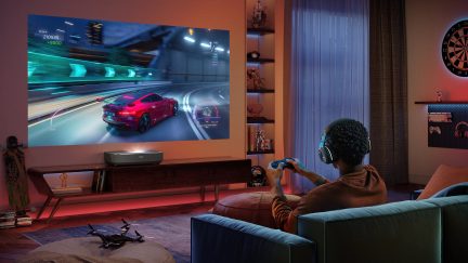 Person auf dem Sofa sitzend, Kopfhörer, Spielcontroller, Wandbildschirm mit Rennspiel, niedriger Schrank, Skateboard, Dartscheibe