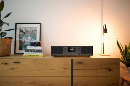 Catena hi-fi nera vista frontalmente, schermo centrale, griglie e pulsanti, piedi inclinati, su mobile in legno chiaro