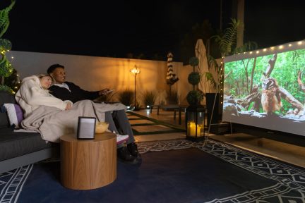 Altoparlante portatile nero compatto su tavolino basso, serata cinema all'aperto con coppia e schermo di proiezione