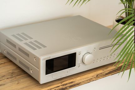 Sølvfarvet audioforstærker i børstet aluminium, set fra højre forside i let fugleperspektiv, med stor sort skærm, bred cirkulær drejeknap, vandret sprække til disk, række af små runde knapper samt ventilationsgitre på toppen og siderne