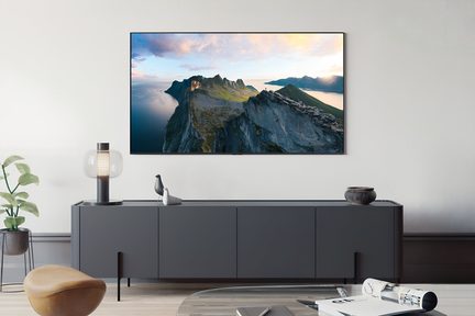 Černý matný televizor s velkým úhlopříčkou a ultra tenkým rámem, hladký materiál pravděpodobně plast nebo kov, pohled zepředu na zeď zobrazující jasnou horskou krajinu