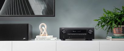 Denon AVC-X3700H sort hjemmebiografsforstærker, set forfra, placeret på et hvidt møbel med trædekorationer og en grøn plante