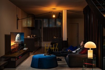 Moderne stue med grå sofa, sofabord, lampe, fladskærms-tv der viser en planet mod en sort baggrund, og en mand der sidder og ser på skærmen