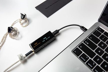 Μικρό εξωτερικό ορθογώνιο μαύρο μεταλλικό audio box με οθόνη και τρία πλευρικά κουμπιά, σε λήψη από ψηλά, συνδεδεμένο με καλώδιο USB-C σε γκρι φορητό υπολογιστή και με στριφτό καλώδιο σε ασημένια in-ear ακουστικά