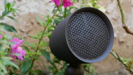 Tmavě hnědý venkovní reproduktor, hladká skříň, perforovaná kulatá kovová mřížka, uchycený na kulovém kloubu, pohled ze tří čtvrtin