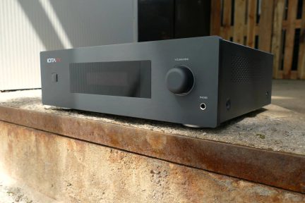 Rektangulärt elektronikhölje i matt svart metall, sedd snett framifrån vänster, front med vitt IOTAVX-logo, centrerat mörkt displayfönster, liten fyrkantig knapp till vänster, stor rund ratt till höger under texten ”VOLUME/MENU” samt hörlursuttag.