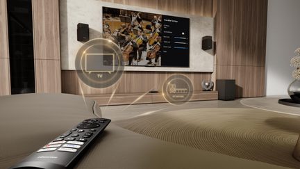 Fugleperspektiv fra en beige lædersofa over en sort plastikfjernbetjening med farvede knapper, foran en medievæg i brunt træfiner med et sort fladskærms-tv, to væghøjttalere og gyldne holografiske ikoner, der angiver TV og soundbar.