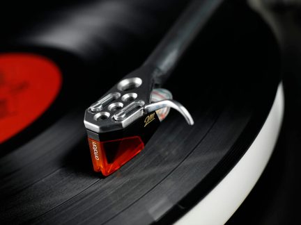 Gros plan d'une cellule phono sur disque vinyle, porte-cellule métallique ajouré, pointe rouge translucide