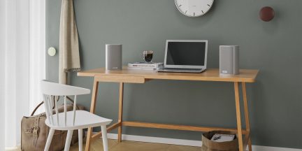 Scrivania in legno chiaro con computer portatile, 2 altoparlanti grigi, tazza di caffè, libri, sedia bianca, borsa in pelle, orologio da parete