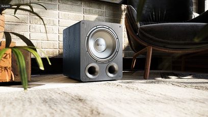 Subwoofer SVS PB-1000 PRO černý jasan, pohled zepředu, umístěný na podlaze na koberci, interiér s dřevěným vybavením, kožené křeslo