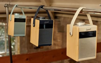 Tre piccole radio portatili rettangolari in bambù naturale, con frontali verde pastello, nero e beige a griglia orizzontale, grande pulsante circolare argentato sulla parte superiore, maniglie abbinate, appese a una rotaia metallica, vista in leggera controplongée