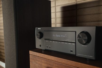 Schwarzer Denon-Heimkinoverstärker auf einem Holzmöbel, Front mit Digitaldisplay und großen runden Drehknöpfen