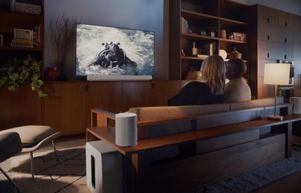 Bílý soundbar Sonos pod TV, subwoofer Sub a reproduktor One v útulném obývacím pokoji s párem na pohovce