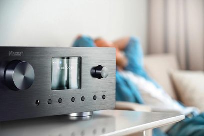 Magnat MC 400 lydsystem i børstet aluminium med blå LCD-skærm, knapper og stik, ved siden af en uskarp liggende person