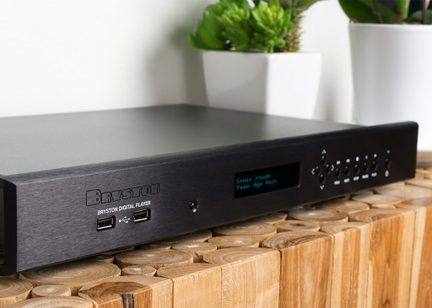 Player audio digital Bryston negru, panou frontal cu ecran și butoane, așezat pe un mobilier din lemn deschis la culoare, cu plante verzi
