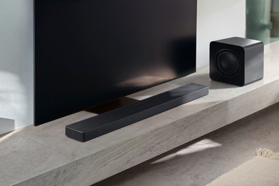 Černý zvukový panel s perforovanou kovovou mřížkou, pohled z levé přední perspektivy pod televizí, doplněný o černý matný subwoofer ve tvaru krychle s kruhovým reproduktorem umístěným vpravo