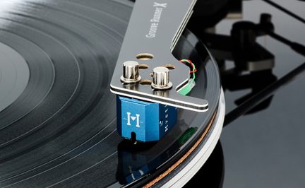 Skråt nærbillede af en blå phono-pickup i anodiseret aluminium, monteret under et udskåret headshell i børstet metal og fastgjort med to kromskruer, placeret på en skinnende sort vinylplade