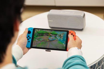 Преносима конзола Nintendo Switch с Joy-Con в синьо и червено, показваща цветна приключенска игра, държана в ръце върху бяла маса