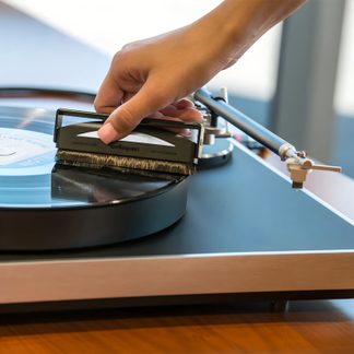 Pagrindinis asmuo laiko stačiakampį šepetį su šereliais ant juodo vinilo disko, padėto ant patefono su matomu tonarmu.