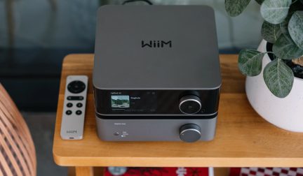 Un dispositivo audio grigio con schermo e pulsanti circolari, un telecomando grigio a sinistra, una pianta in vaso a destra