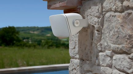 Diffusore da esterno bianco su supporto a parete, fissato a un muro in pietra, vista laterale