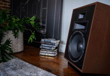 Hnědá dřevěná hi-fi reproduktorová skříň s černým čelem, velkým wooferem a dvěma výškovými reproduktory, pohled ze tří čtvrtin na parkety v interiéru