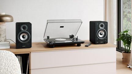 Black vinyl turntable with a raised transparent lid, two black wood bookshelf speakers, on a light wood furniture piece