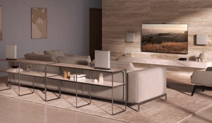 Modernus salonas su pilka sofa, metaliniu konsoliniu stalu, lentynomis, sieniniu televizoriumi, garsiakalbiais, smėlio spalvos kilimu, akmenine siena.